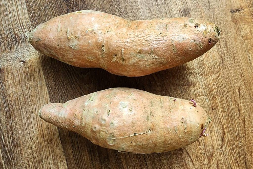 two sweet potatoes on a wood background. they each have a couple tiny sprouts on the growing end