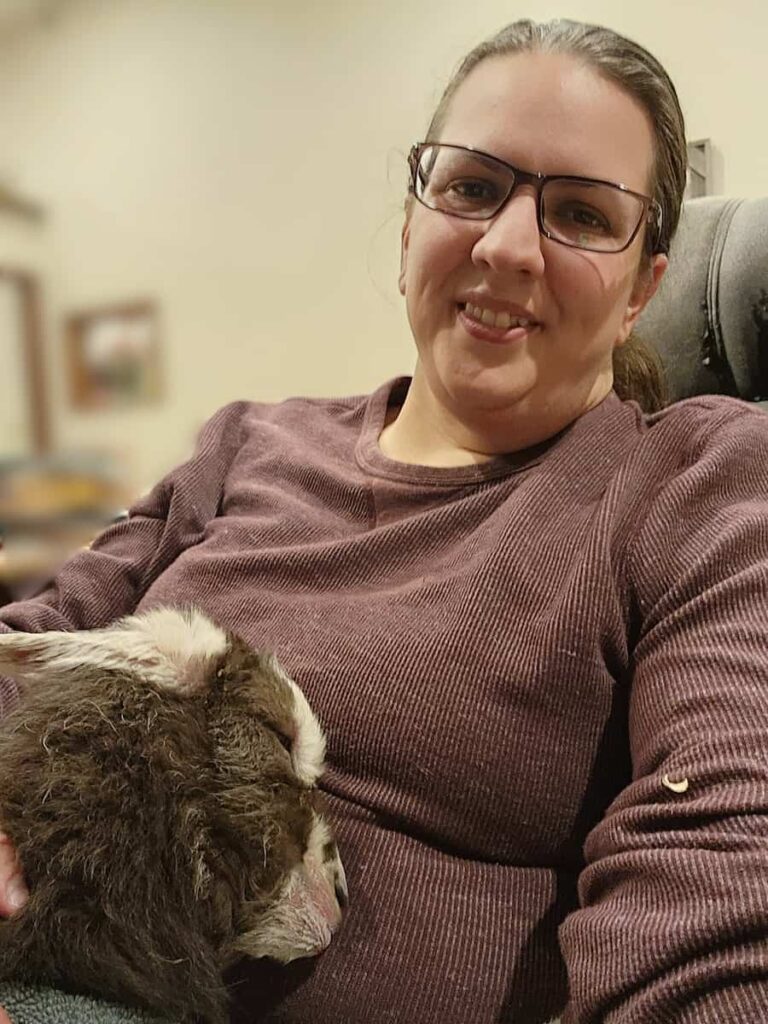 A woman wearing glasses and a maroon sweater smiles while a small animal, possibly a baby goat or lamb, rests its head on her chest. The background is softly blurred indoors.